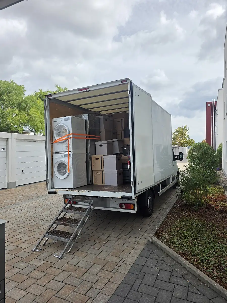Beladener Koffer-LKW mit Stahltreppe am Heck: Waschmaschine und Trockner mit Spanngurt gesichert, dahinter gestapelte Umzugskartons